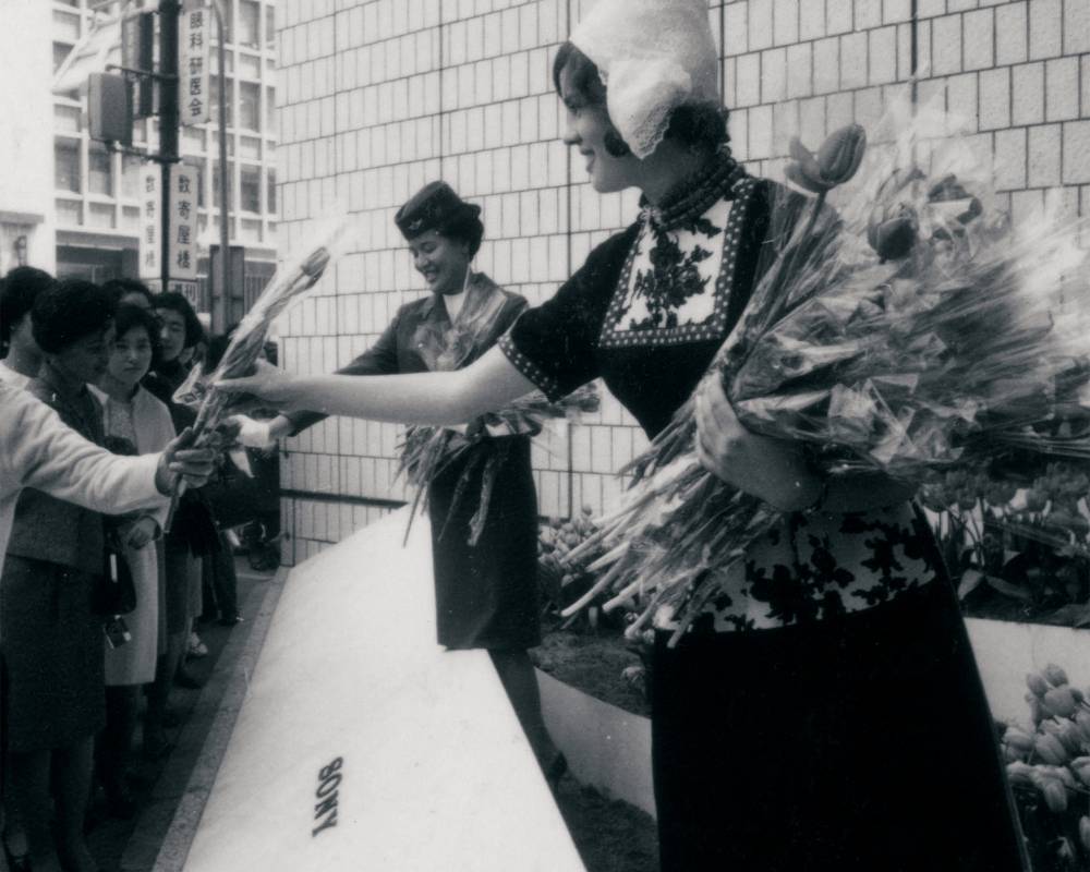 An event (black-and-white). Women in traditional Dutch dress and flight attendants hand out tulips at Sony Square (outdoor event space).
