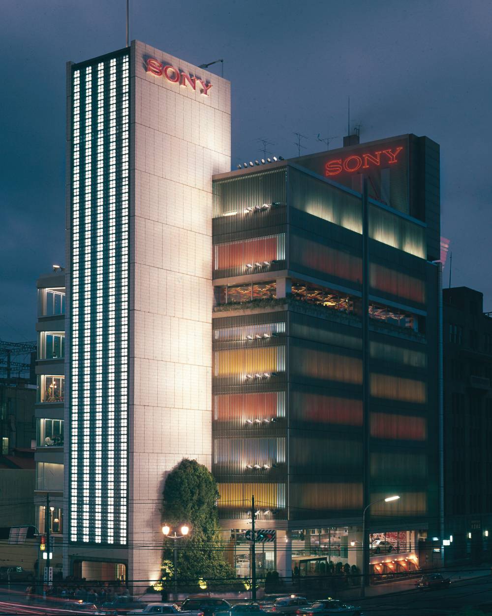 Sony Building exterior. Panoramic view of the Sony Building at night, beyond Sukiyabashi Crossing. A three-story-high Japanese andromeda tree has been planted in Sony Square (outdoor event space) in front facing the intersection.