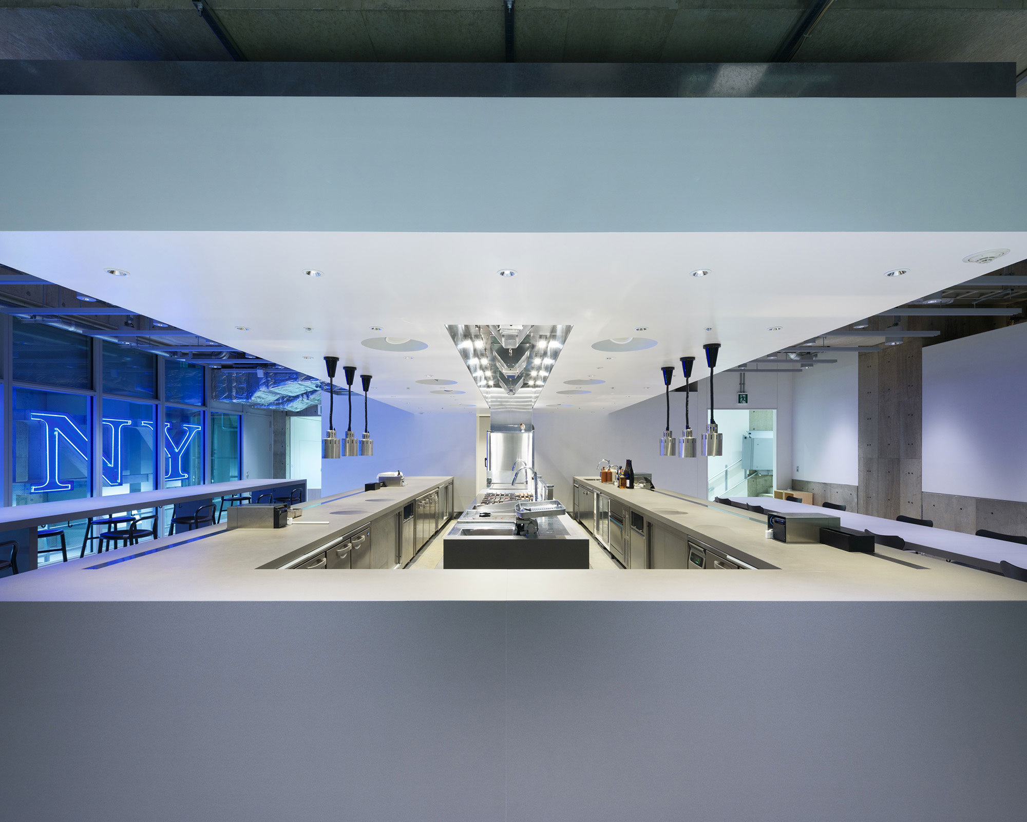 An interior photo of a restaurant. The kitchen is in the center, with high tables on the left and low tables on the right.