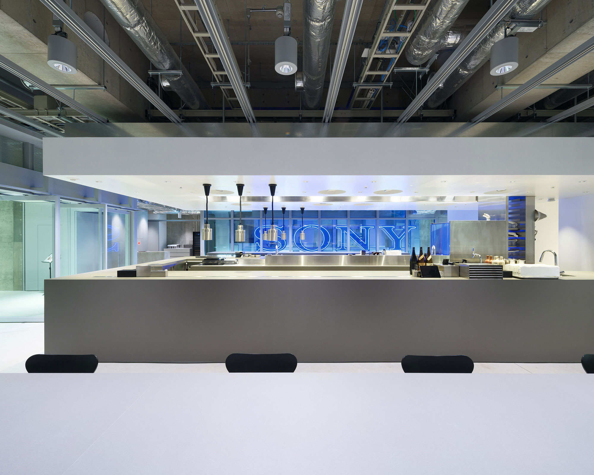 An interior photo of a restaurant. Low tables are in the foreground, with the kitchen and a Sony neon sign in the background.