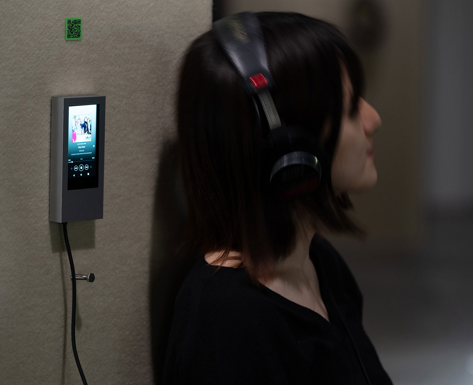 Exhibit. A visitor wears headphones and listens to music from a device mounted on a pillar.