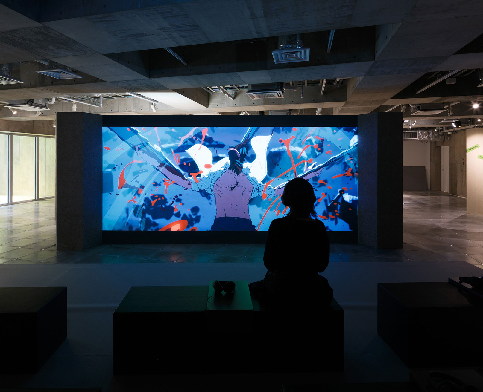 Exhibit. A visitor sits on a bench, watching animated footage projected on a large screen.