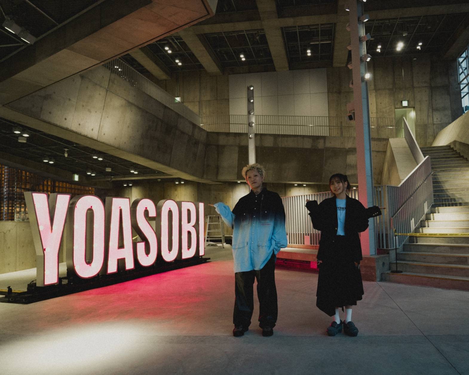 Interior. The two members of YOASOBI posing in front of their YOASOBI neon sign.