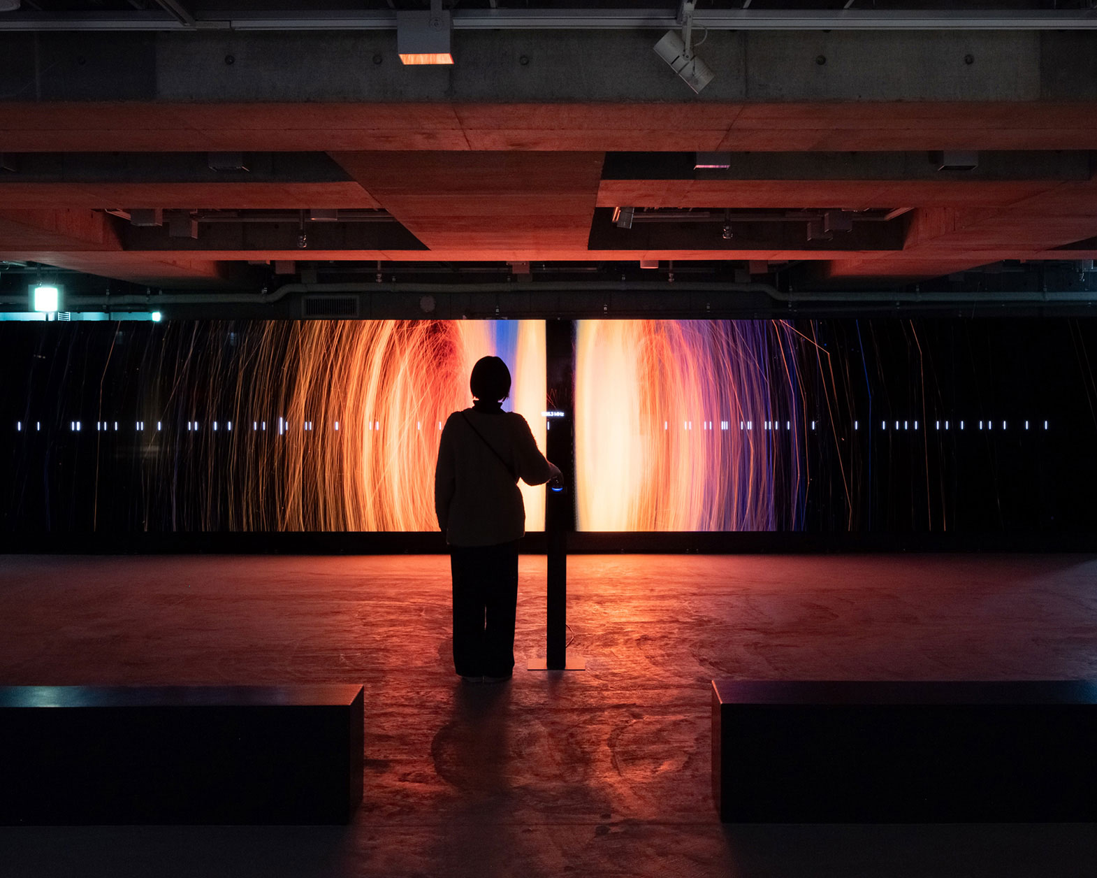 At the back of the dimly lit space, there is a large display showing red and orange visuals, with a person standing in front of it.