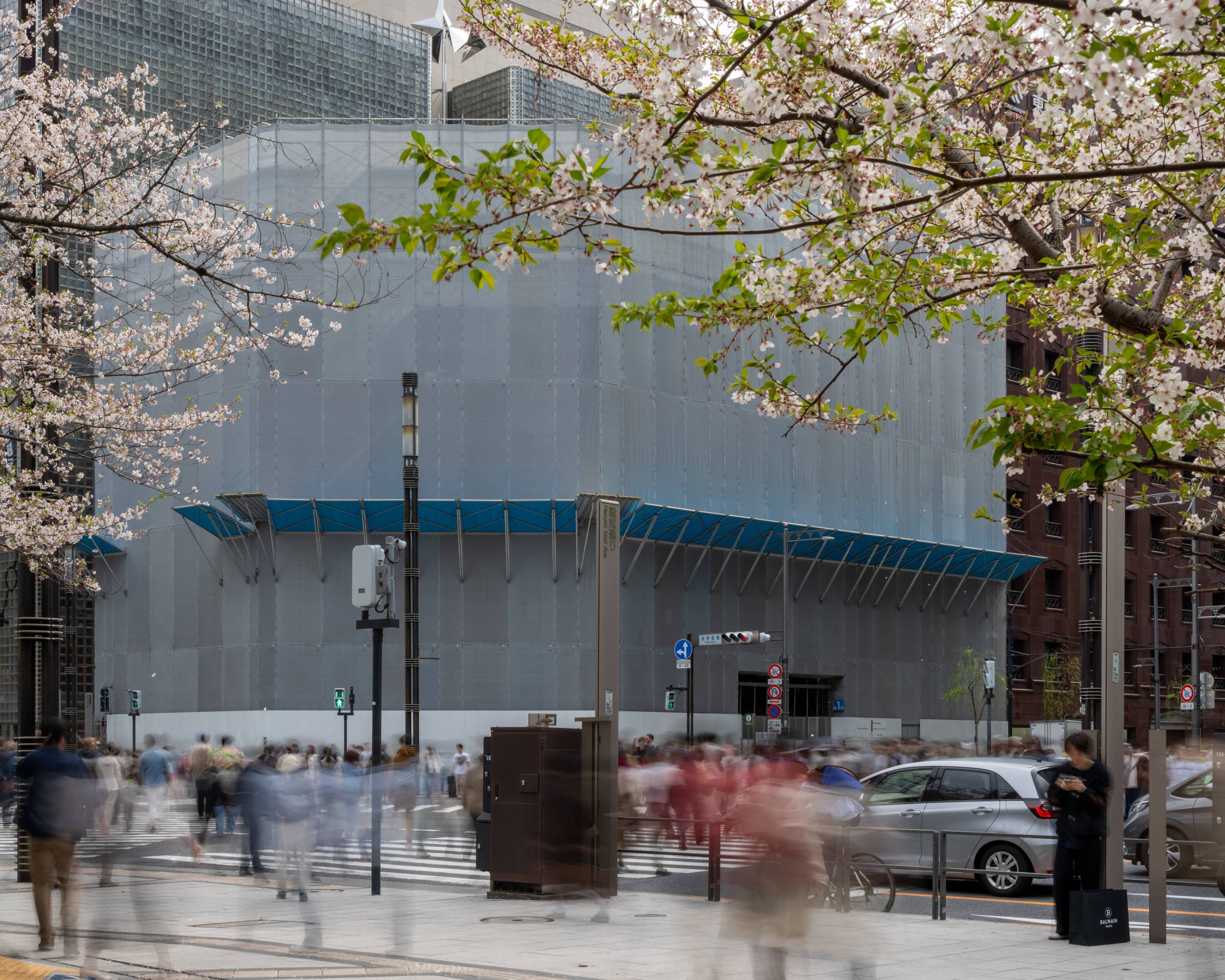 Exterior. People walk to and fro across Sukiyabashi Crossing, between Ginza Sony Park on the far side, covered in its temporary construction fence up to the height of the tower, and the cherry tree in the foreground, with its young leaves.