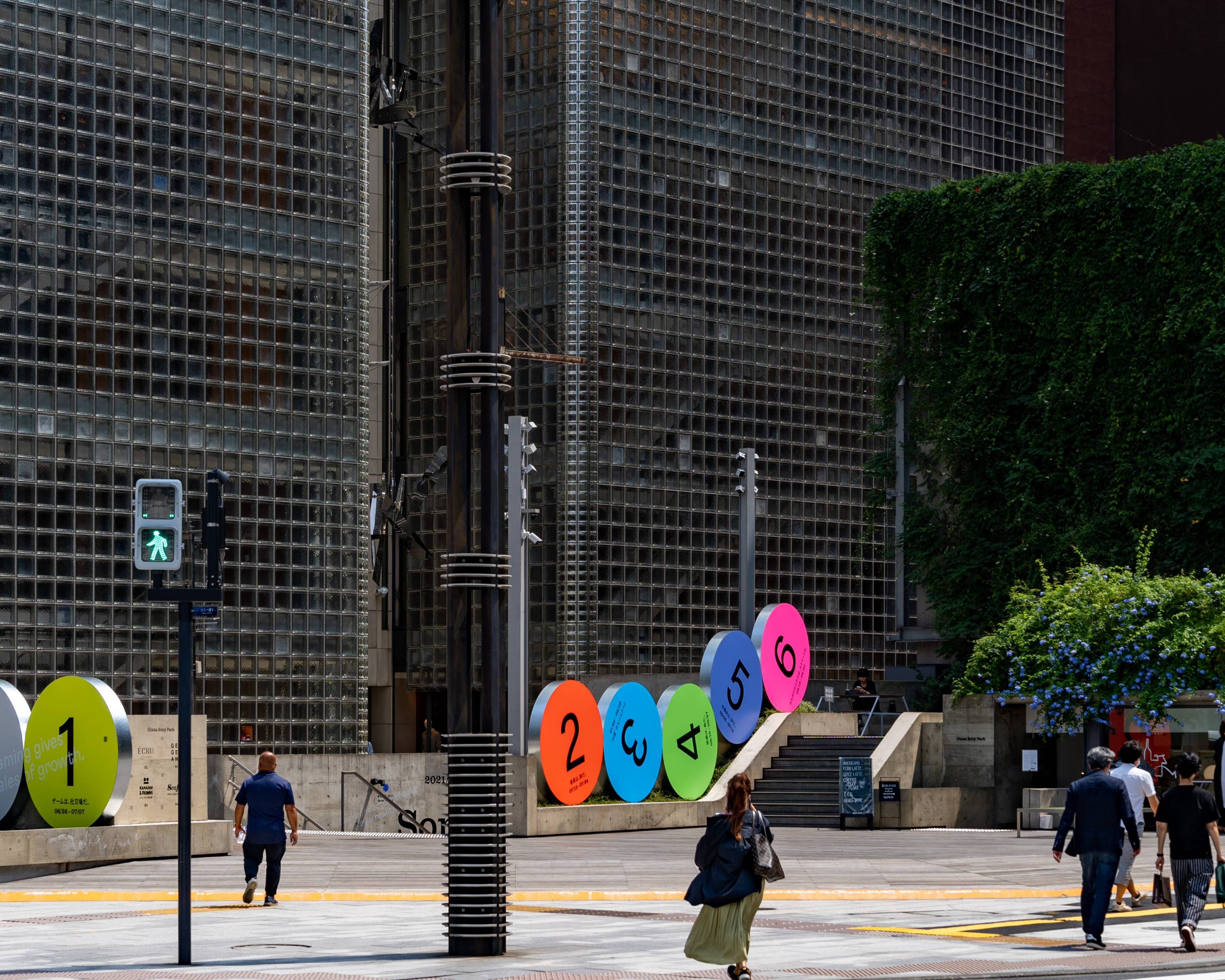 Exterior. Six round art objects, labeled “1” to “6” respectively, are lined up in a row on the ground floor. People walk on the sidewalk next to it.