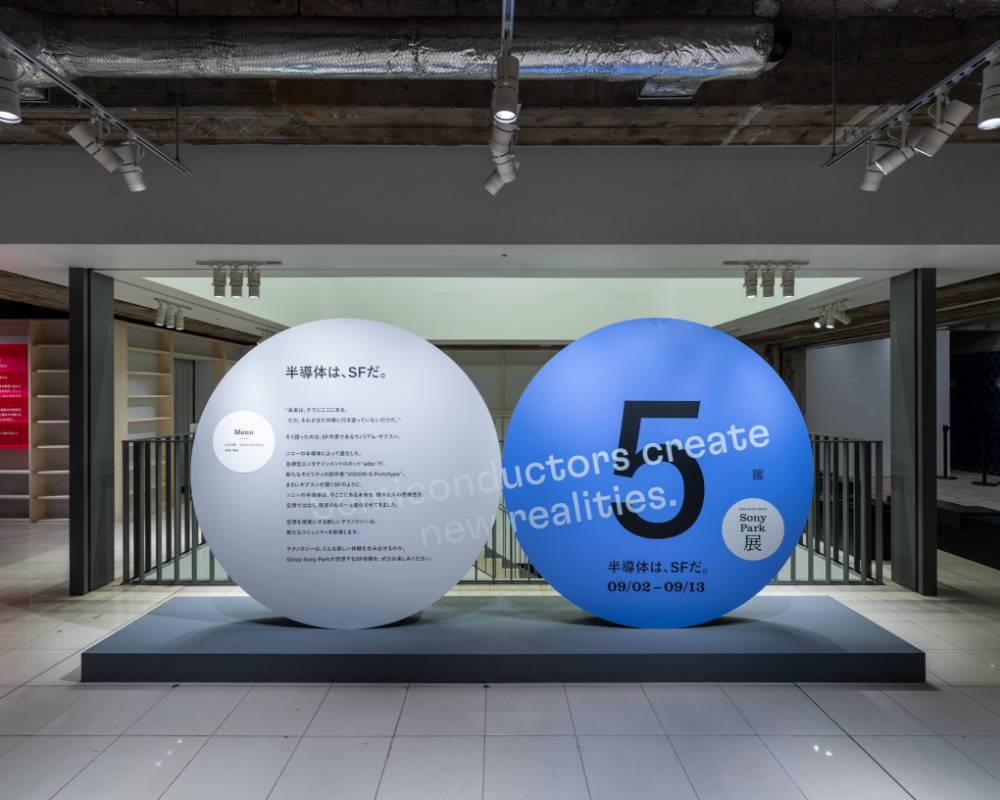 Exhibit. A round gray art object with the exhibition statement, and a blue version of the same object labeled “5,” are displayed side by side at the entrance to the venue on the second basement floor.