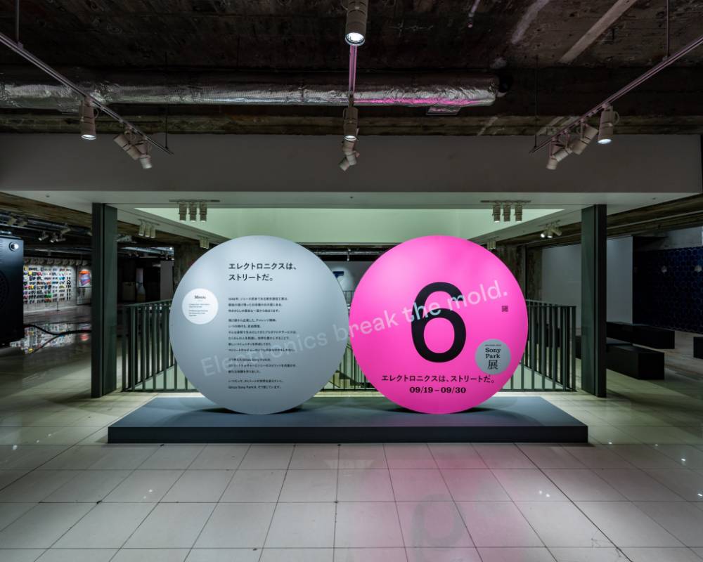 Exhibit. A round gray art object with the exhibition statement, and a pink version of the same object labeled “6,” are displayed side by side at the entrance to the venue on the second basement floor.