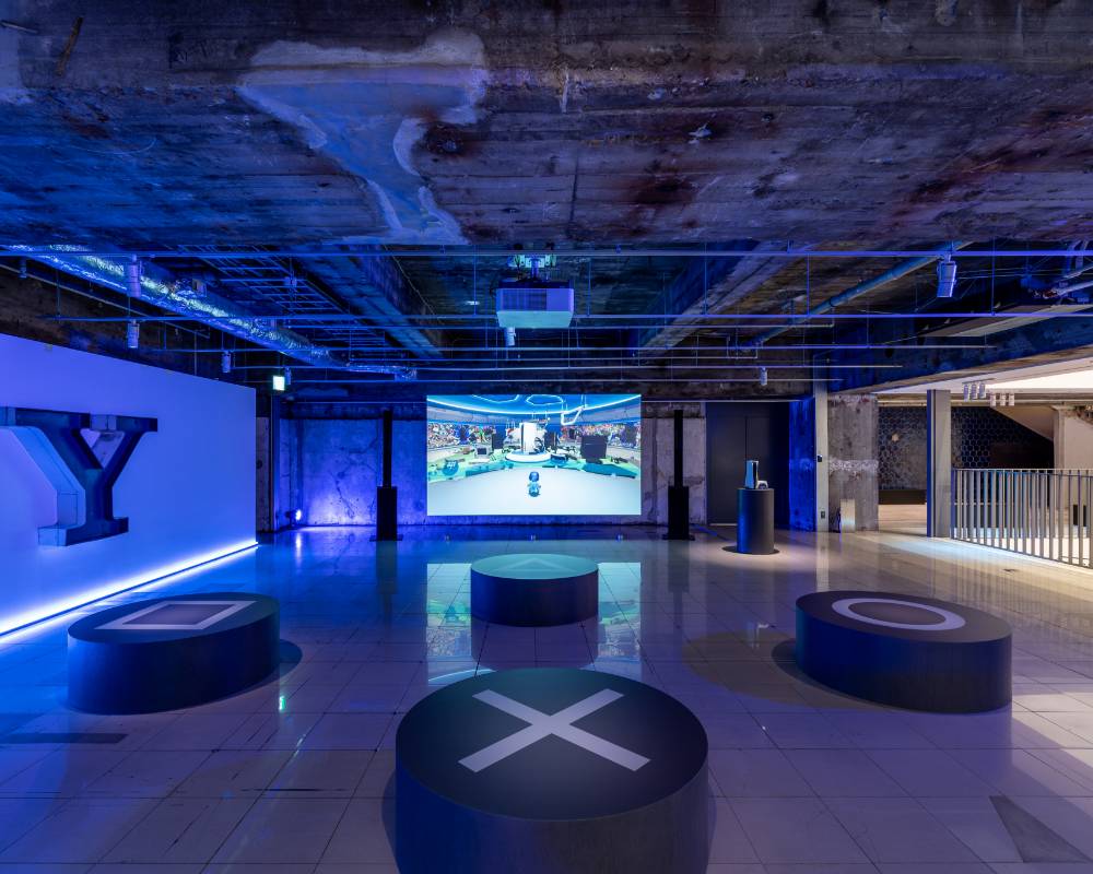 Exhibit. The space has been lit up in blue lighting; a video game screen has been projected onto the back wall, and in front of it, are benches labeled with a square, triangle, circle, and cross mark.