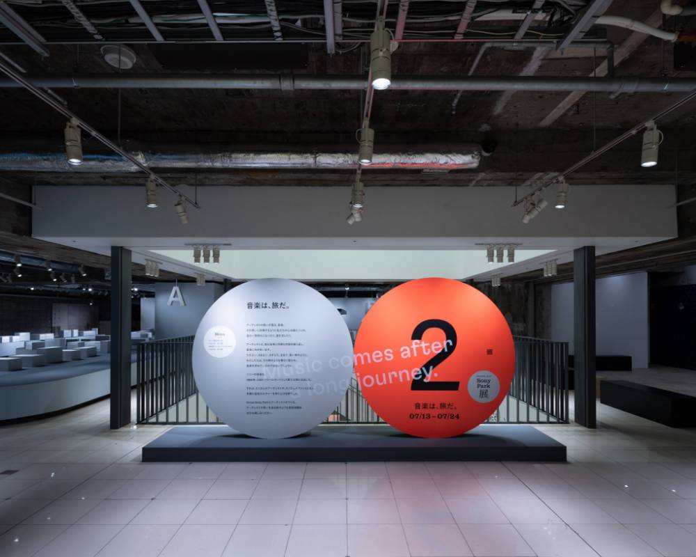 Exhibit. A round gray art object with the exhibition statement, and an orange version of the same object labeled “2,” are displayed side by side at the entrance to the venue on the second basement floor.