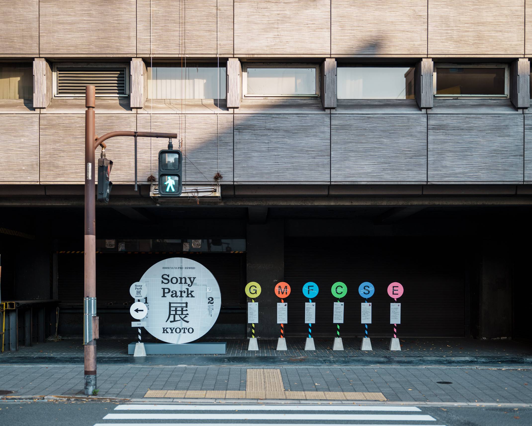 Exterior. Outside, there is a sign with the main visual of the exhibition in a circle, and six signs designed to look like bus stops, lined up in a row.