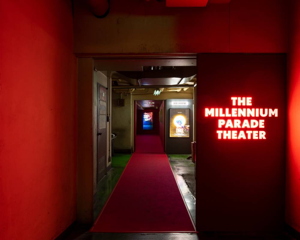 Exhibit. A red carpet has been laid out in the corridor leading to the theater in the back, and an LED “THE MILLENNIUM PARADE THEATER” sign glows red on the entrance door, which is surrounded by deep red walls.