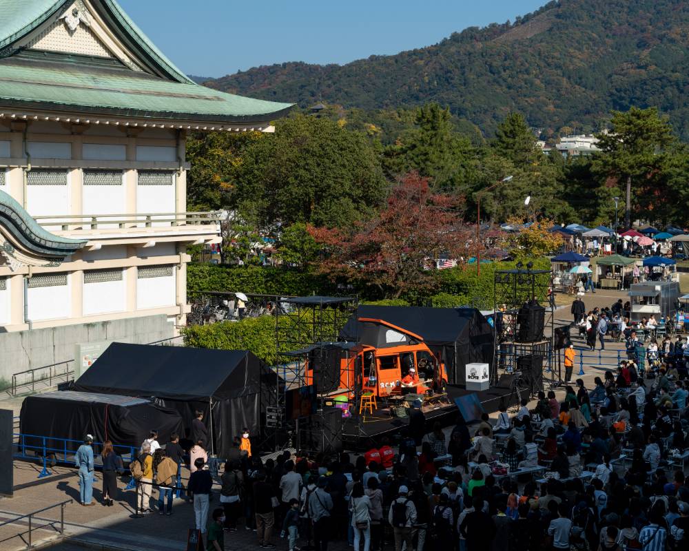 Live performance. A large crowd of people has gathered for a live performance by Tamio Okuda, in front of an orange camping car parked at ROHM Square outside of ROHM Theatre Kyoto.