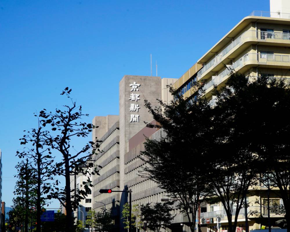 Exterior. Beyond the trees lining the street is a building with the company name “Kyoto Shimbun” displayed on it.