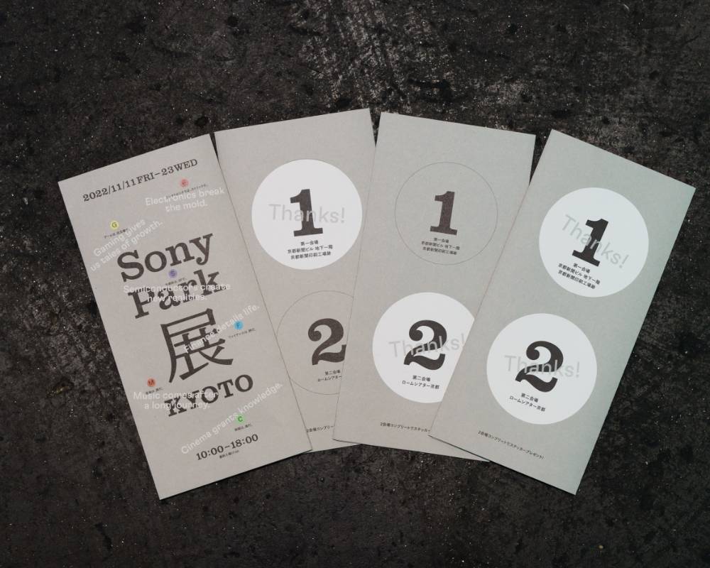Photo of leaflet. The cover features the main visual of the exhibition, and on the back are stickers labeled “1” and “2.”