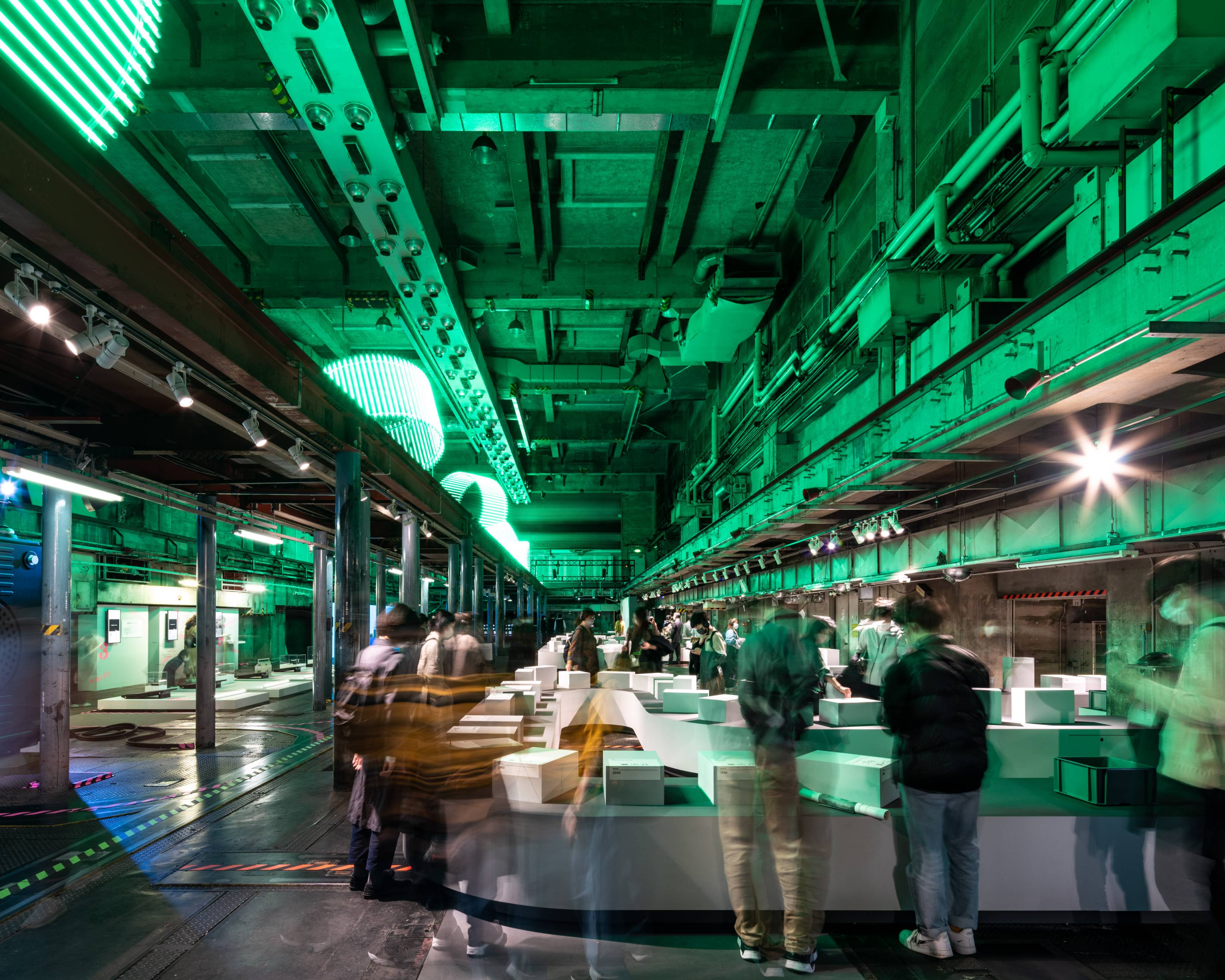 Exhibit. People gather in the exhibition in the underground space, which is lit green by the art object glowing green on the upper level.