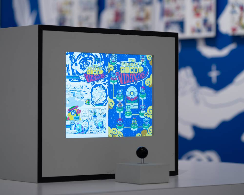 Exhibit item. A monitor shows a page from the manga The Pilgrim’s Guide to Video Games, and in front of it is a game controller stick.