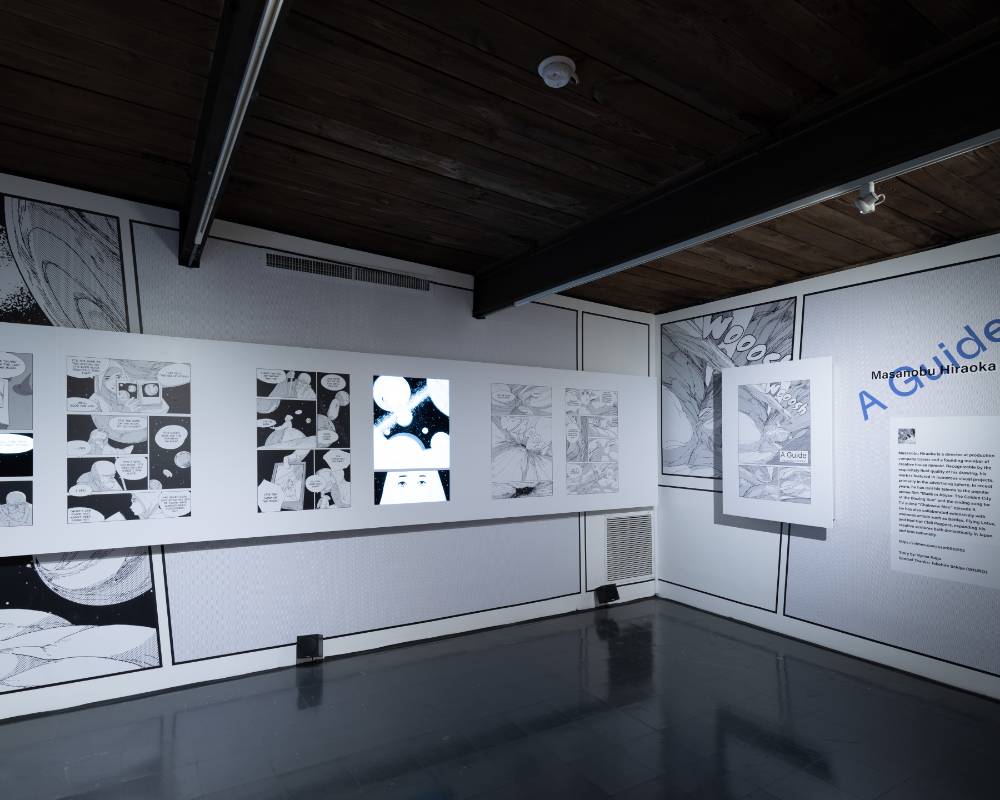 Exhibit. Individual pages from the manga A Guide are exhibited on the wall, and videos comprised of panels from the manga play on monitors embedded here and there within the space.