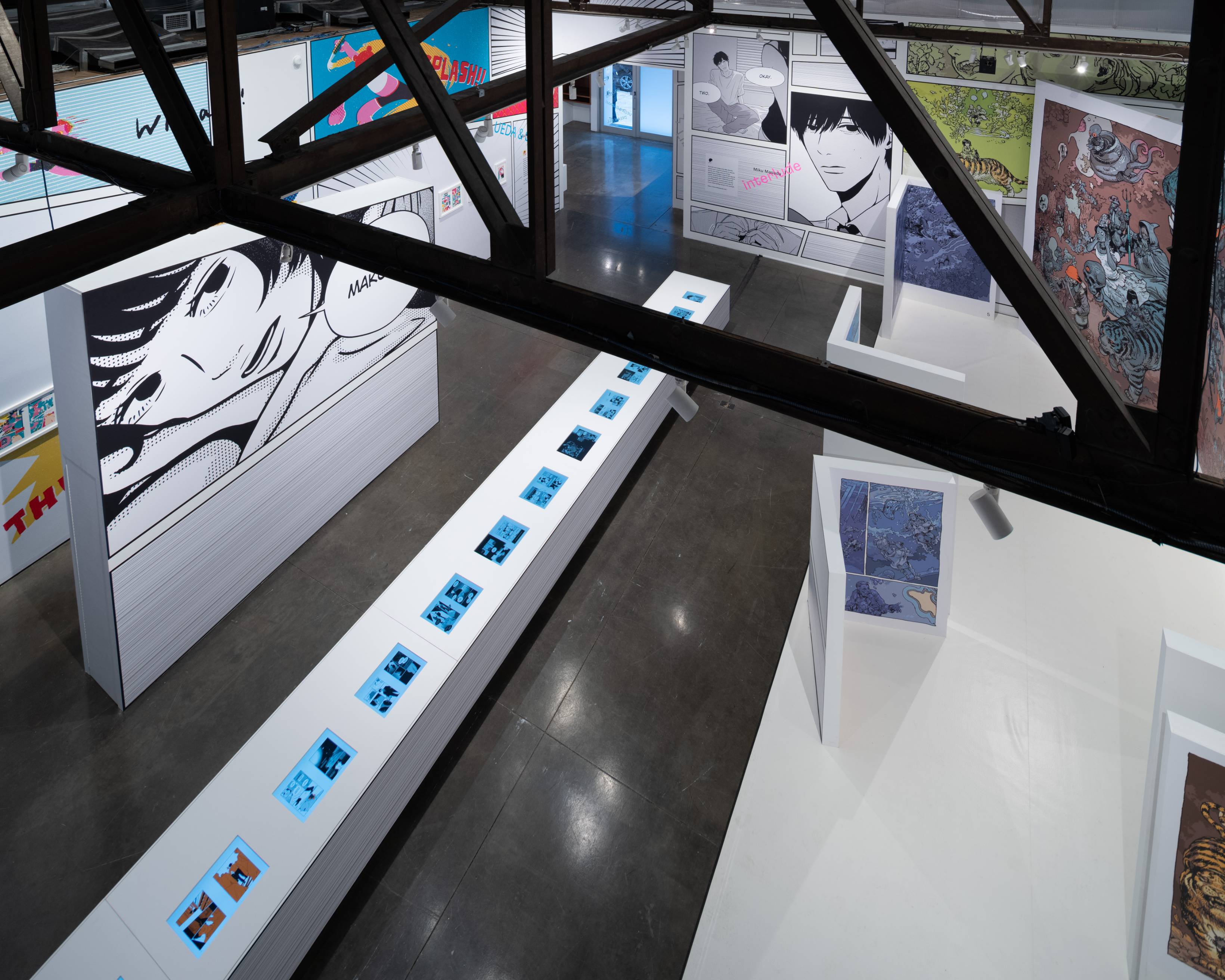 Exhibit. A bird’s eye view of the first floor of the venue shows walls and partitions with large manga exhibits, and a long table embedded with numerous monitors.
