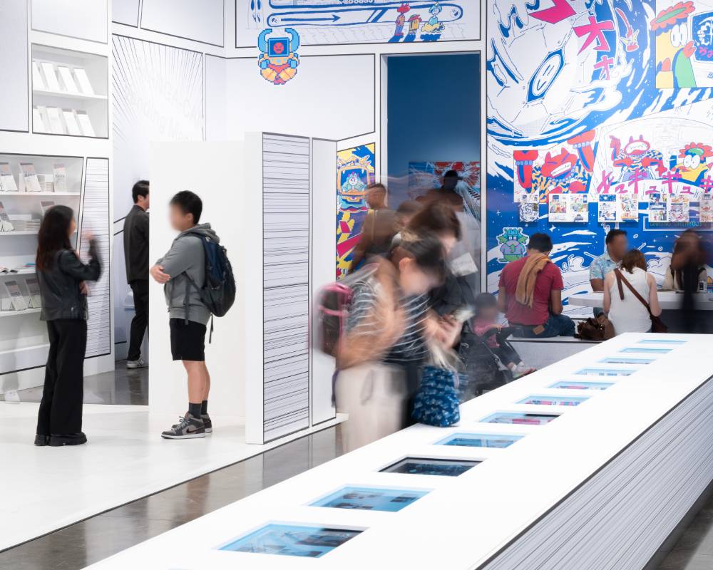 A hands-on experience. Visitors enjoy manga in various different ways, reading manga on the monitors embedded in the table, sitting on a bench reading physical manga, and more.