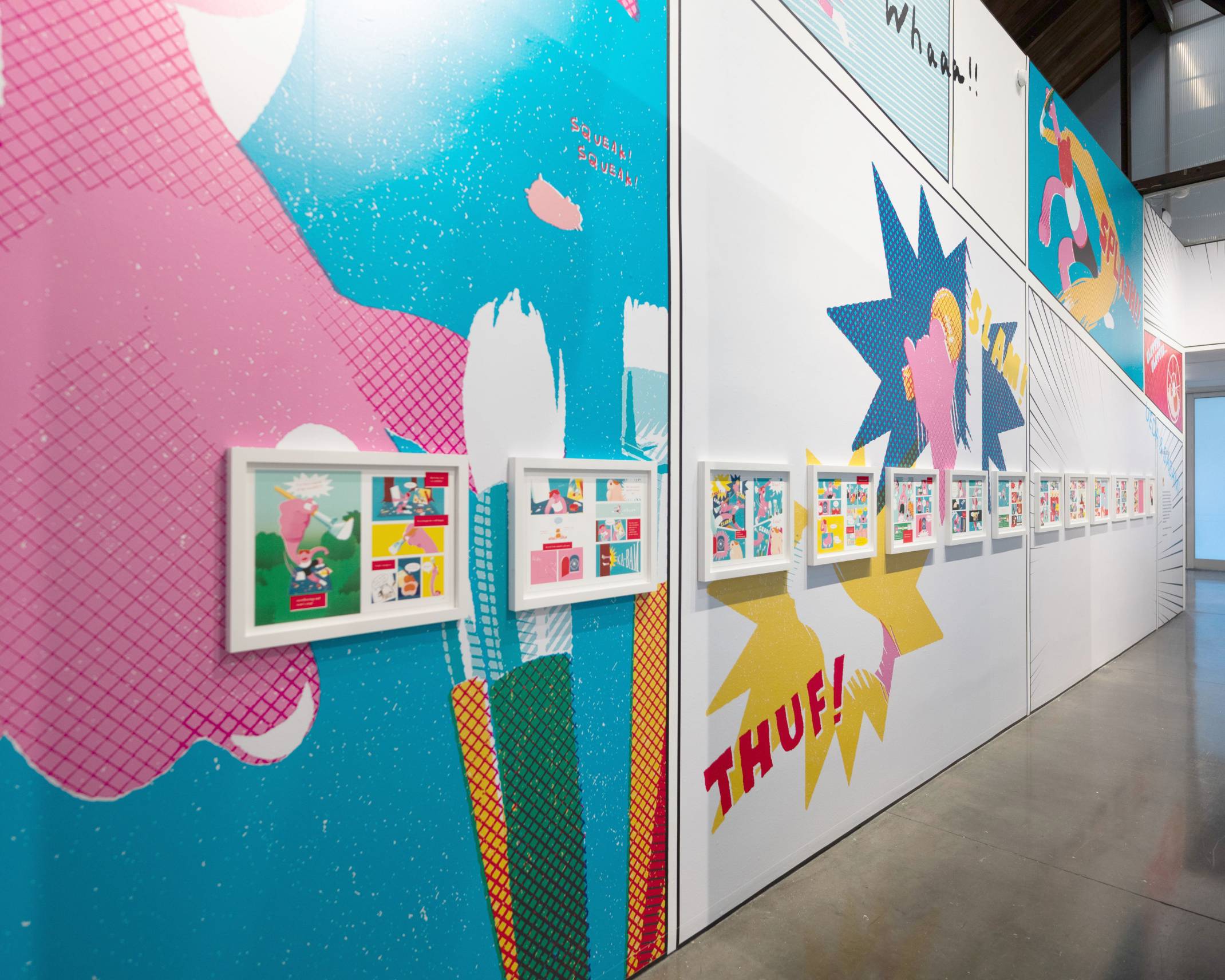 Exhibit. A number of two-page spreads from the manga UEDA & SASAMI, each in a white wooden frame, are exhibited on the wall, which has been decorated with prominent, colorful graphics.