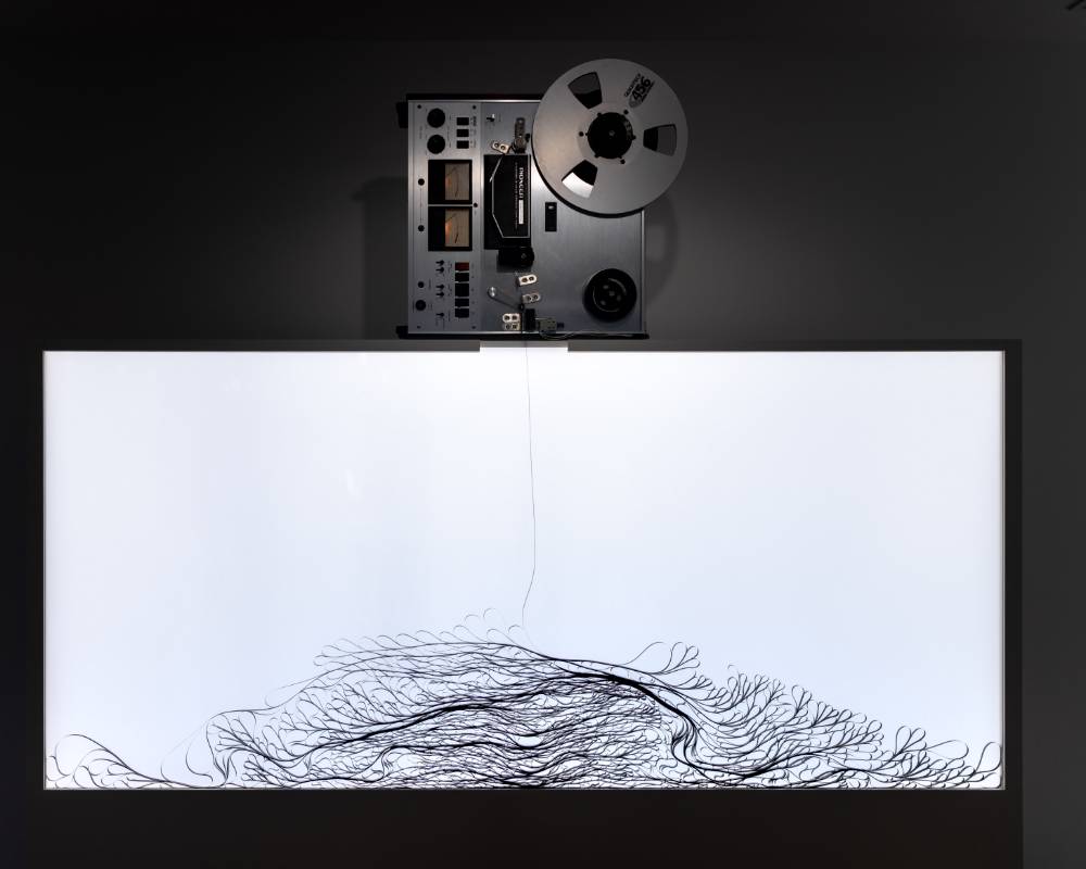 Exhibit. The tape that falls from the reel-to-reel tape recorder folds over and stacks on top of itself inside of the container.