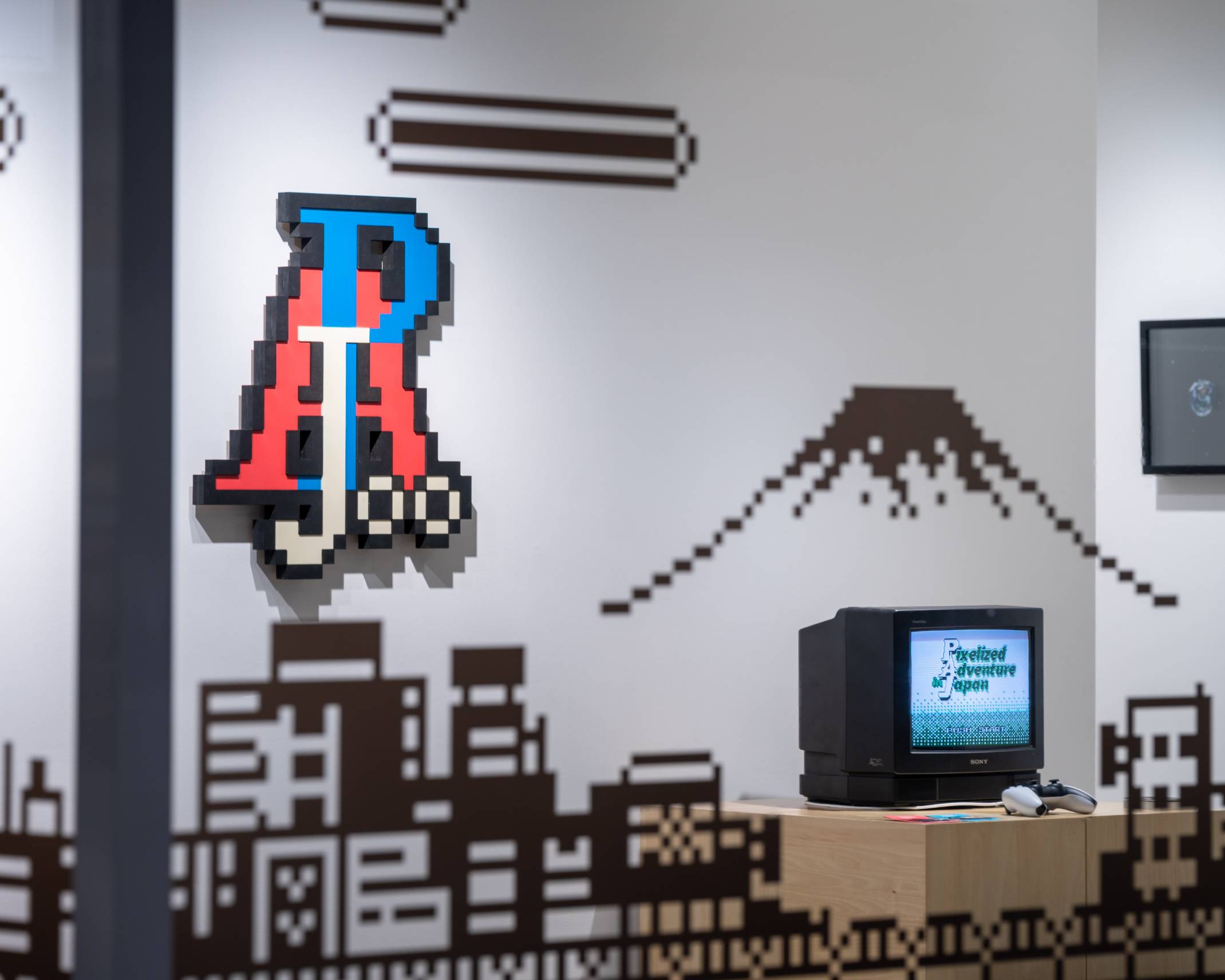 Exhibit. The wall is decorated with pixel art, and placed on a platform is a CRT TV showing a game screen, and a controller.