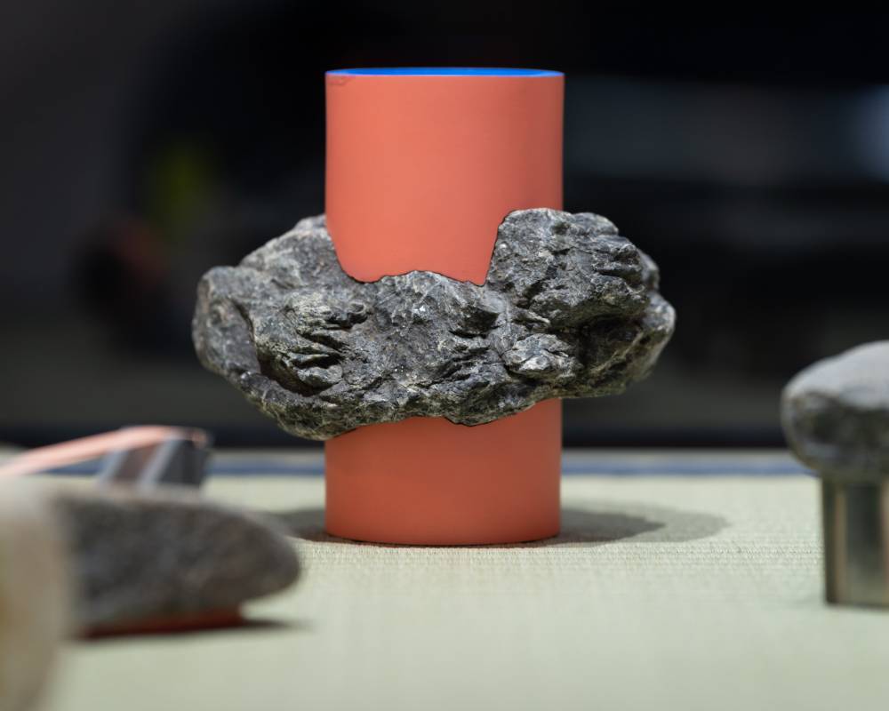 Exhibit item. An art object, comprised of a brick-colored cylinder with stone seemingly wrapped around it, is displayed.