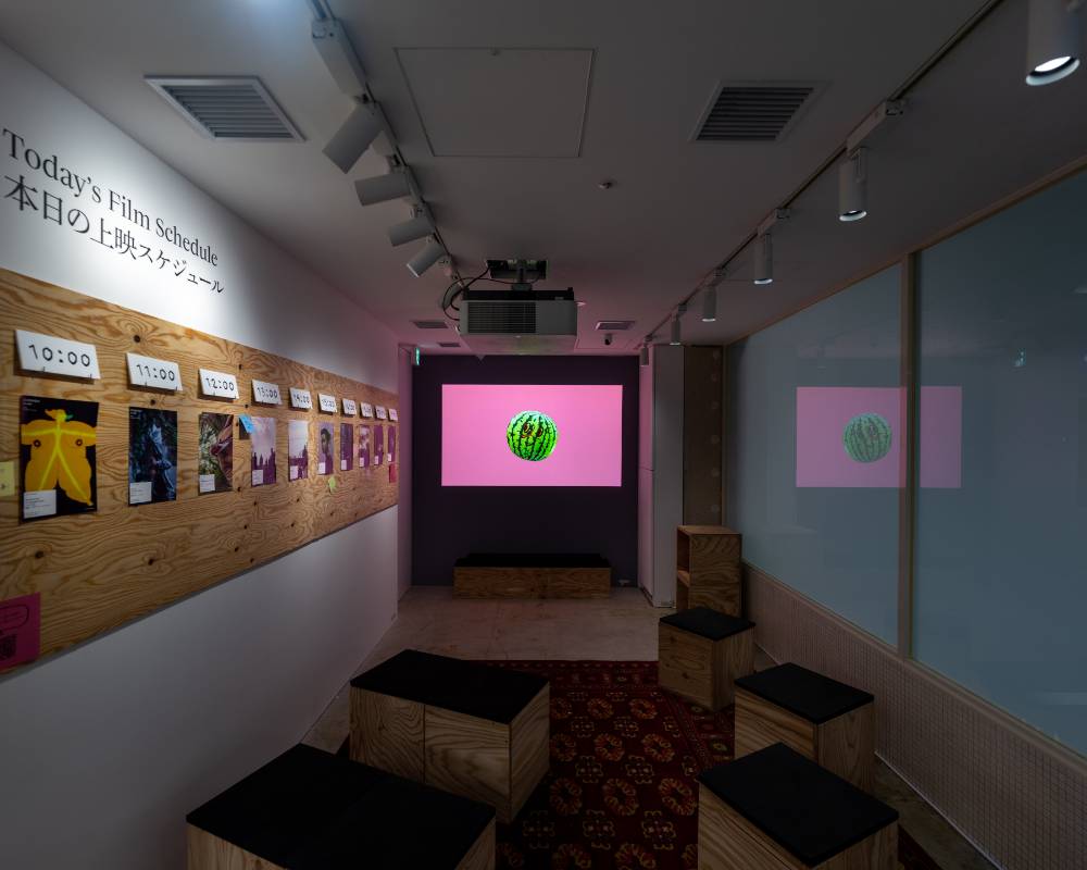 Interior. A video has been projected on the wall on the far side of the exhibition space, with several stools placed in front of it.
