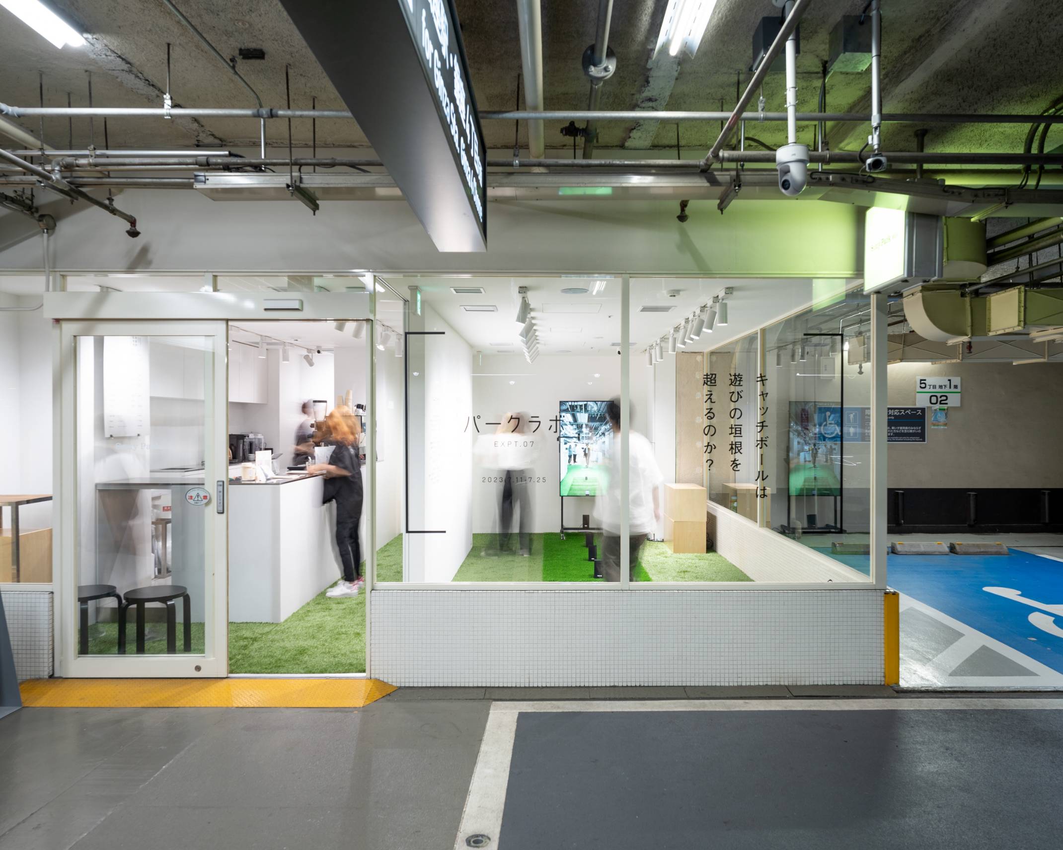 Exterior. The floor of the exhibition space is covered in green artificial turf, and in the back of the space is a vertical monitor.