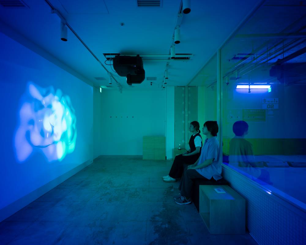A hands-on experience. Two visitors with devices on their heads sit in the space, watching a video projected onto the wall.