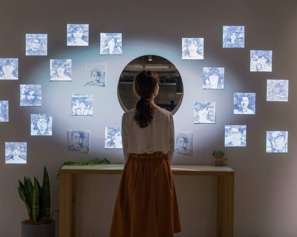 A hands-on experience. A visitor stands in front of a round mirror placed in the center of a wall, on which numerous faces have been projected.
