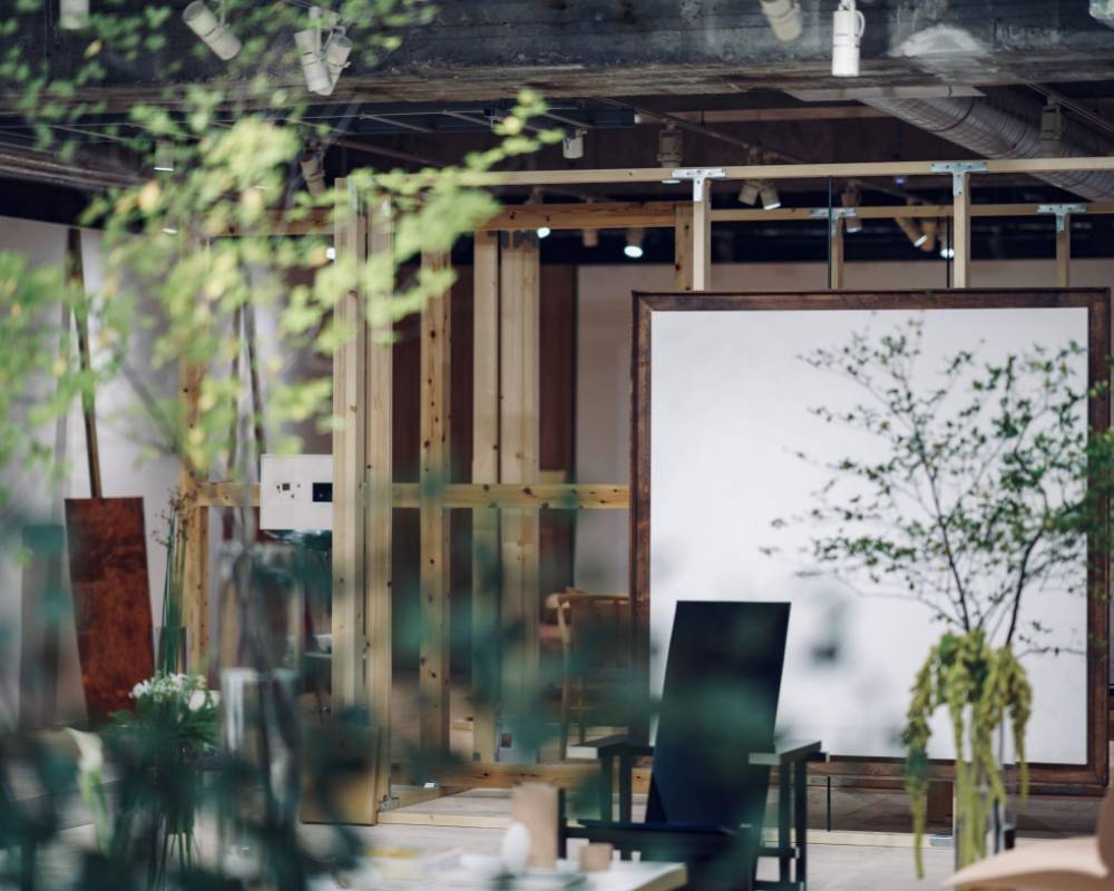 Exhibit. A large white painting and multiple chairs, including a black chair, can be seen exhibited past the branches of plants and trees.