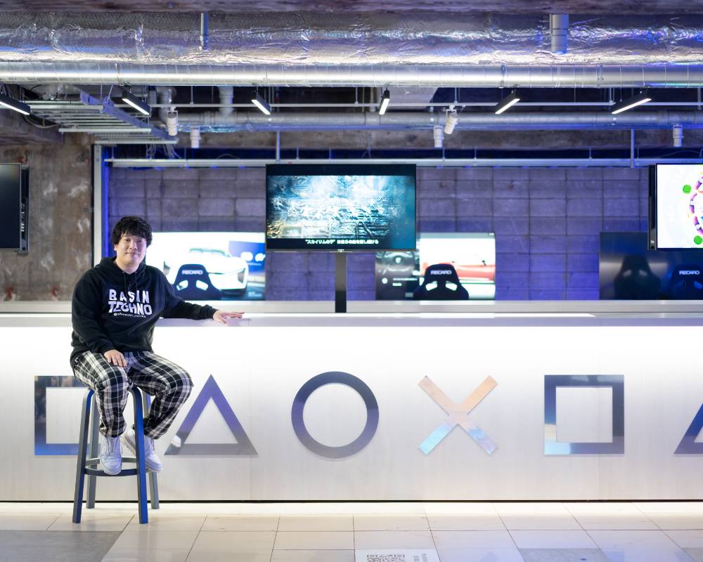 Exhibit. okazakitaiiku sits on a chair in front of a large counter featuring square, triangle, circle, and cross marks.