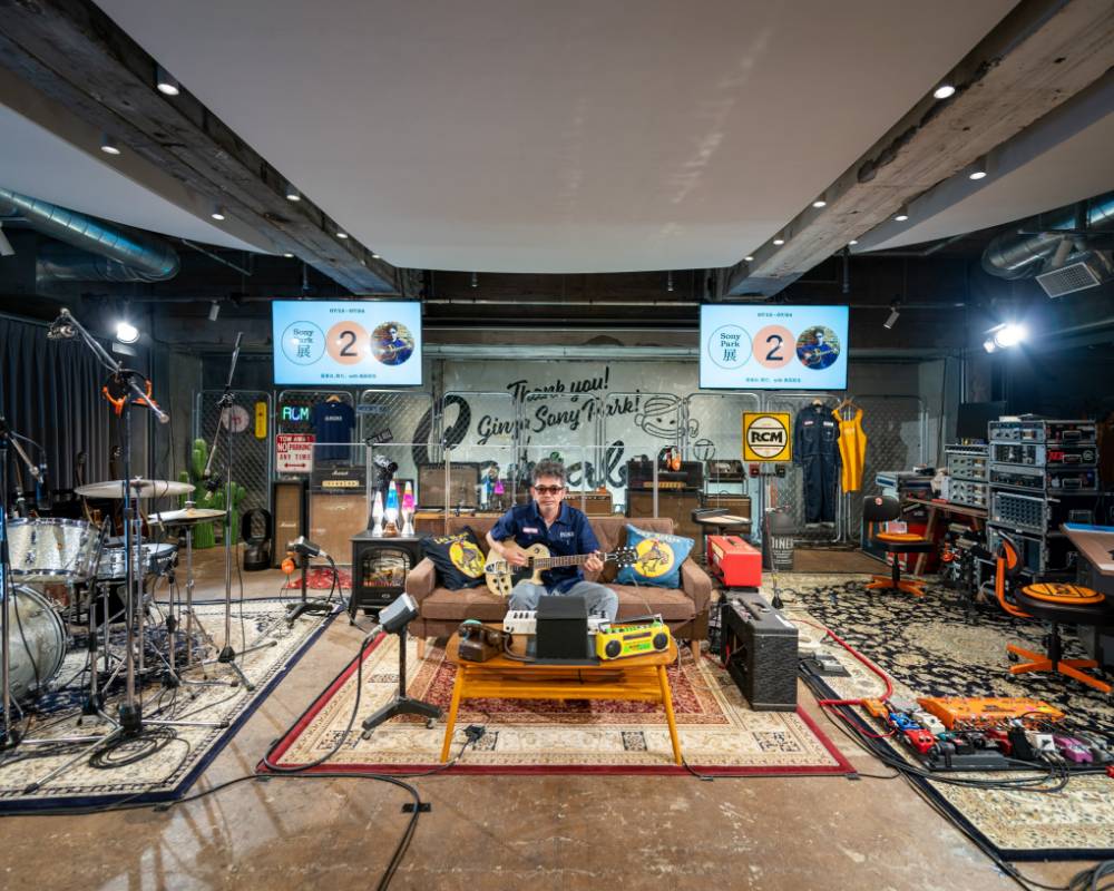 Exhibit/live performance. Tamio Okuda sits holding a guitar on a sofa in the middle of the space, which has been designed to recreate a home recording studio-style space.