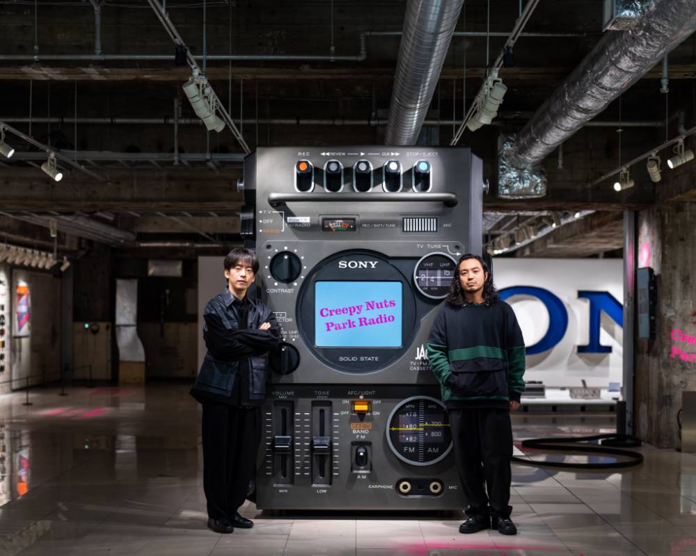 Exhibit. The two members of Creepy Nuts pose in front of the giant radio cassette recorder art object with its integrated TV.