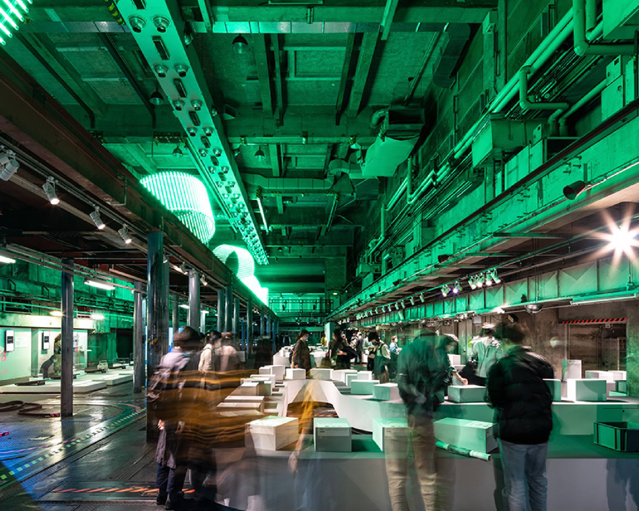 Exhibit. People gather in the exhibition in the underground space, which is lit green by the art object glowing green on the upper level.