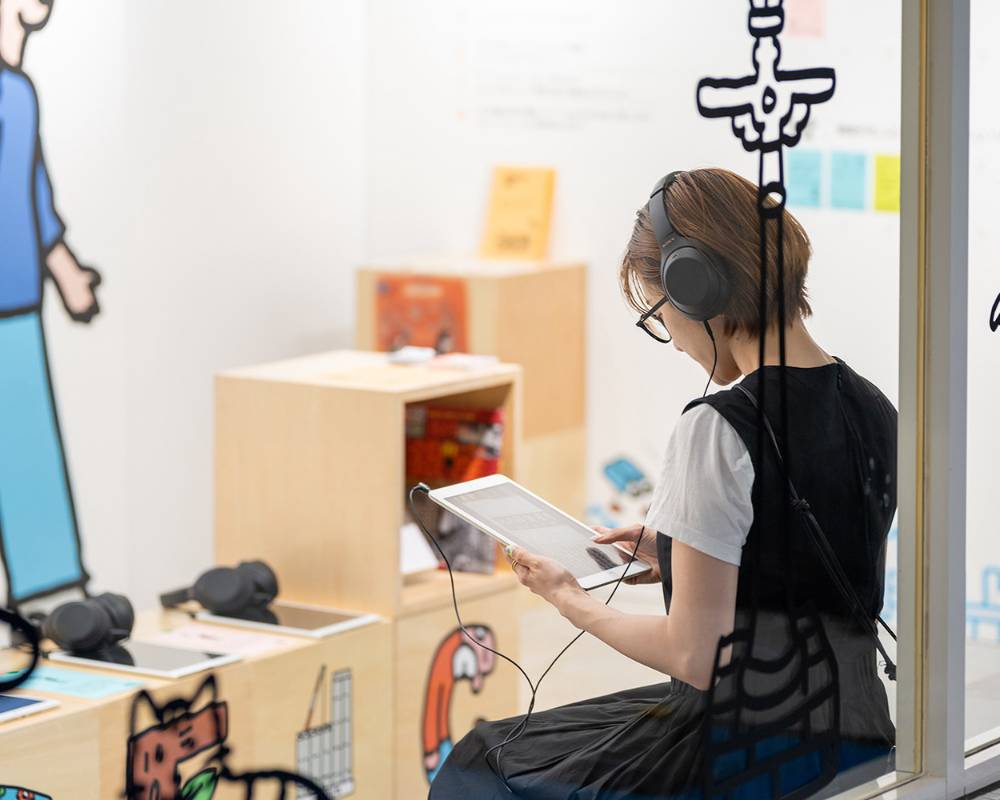A hands-on experience. A visitor sits on a bench with headphones on, gazing at a tablet.