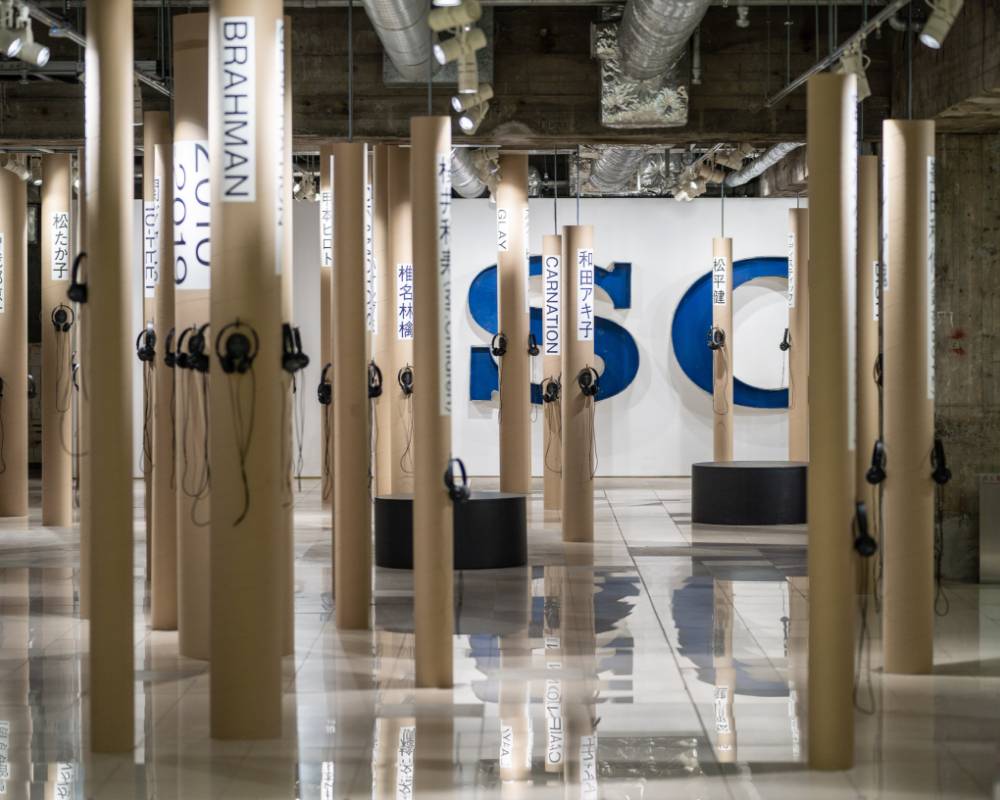 Exhibit. Numerous paper tubes with artist names written on them are stood up in the underground space like trees in a forest, each with a pair of headphones on them.