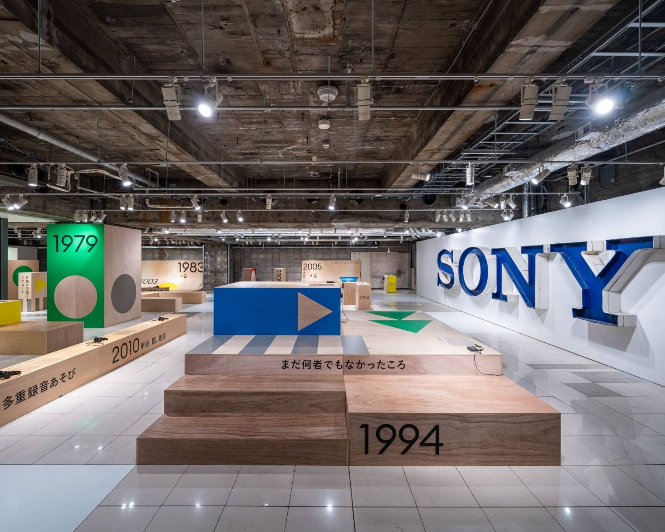 Exhibit. The basement venue, with its large “SONY” sign on the right wall, features wooden display stands assembled to look like playground equipment and labeled with years, on which are displayed WALKMAN models from those years.
