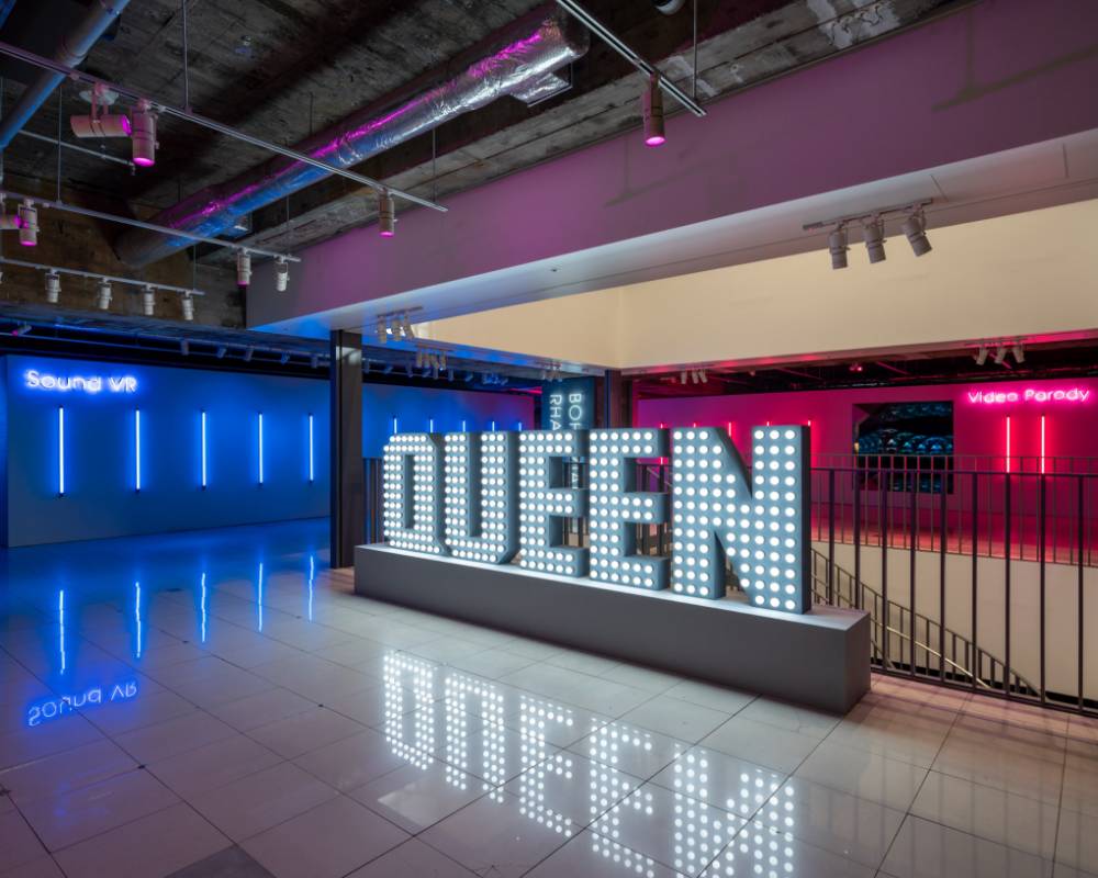 Venue. Electric lights forming the word “QUEEN” are displayed in a space lit with blue and pink LED lights.
