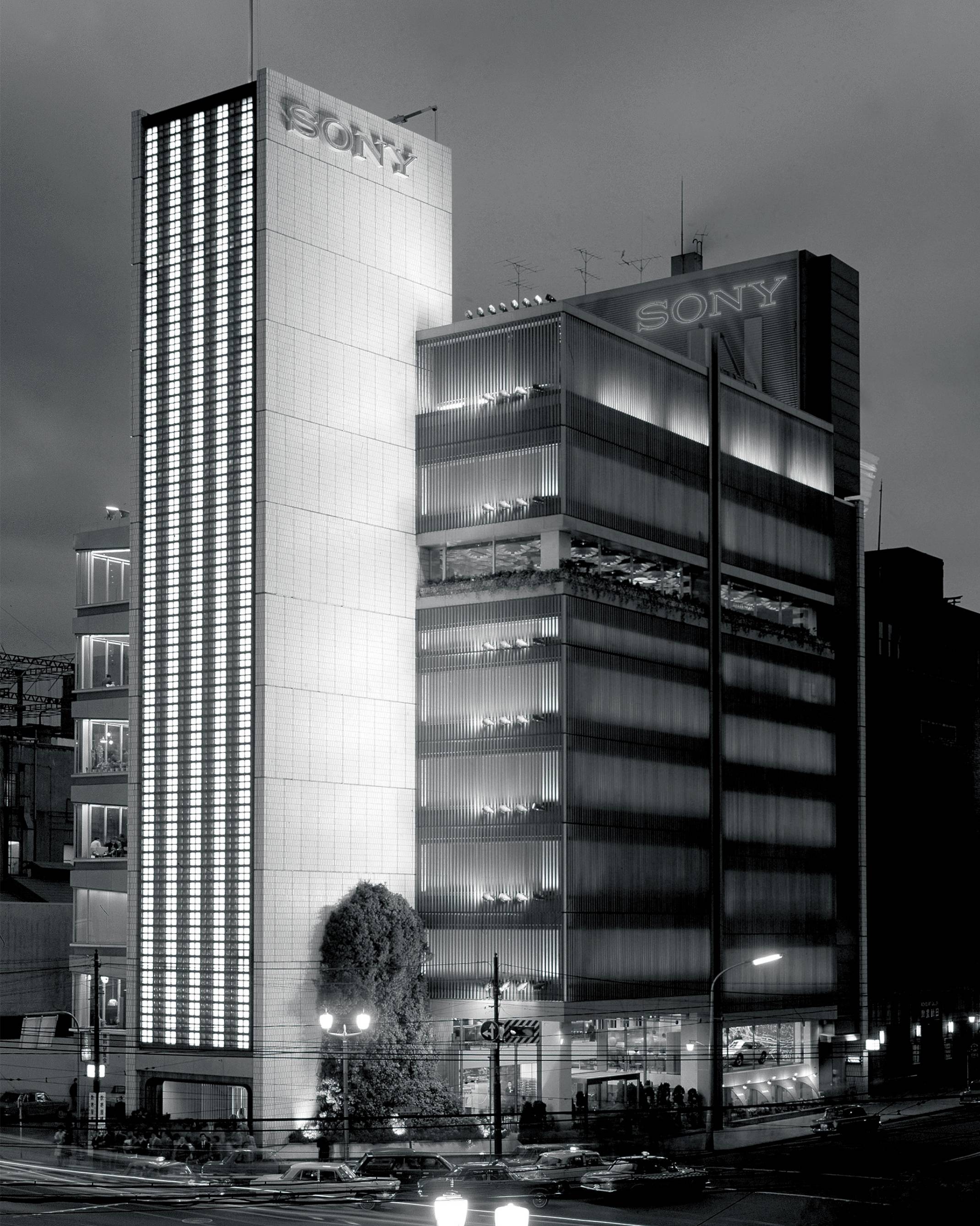 Exterior (black-and-white). Panoramic view of the Sony Building at night, beyond Sukiyabashi Crossing. A three-story-high Japanese andromeda tree has been planted in Sony Square (outdoor event space), which is in the foreground and faces the intersection.