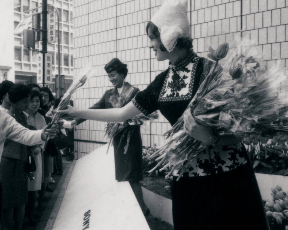 Event (black-and-white). A woman in traditional Dutch dress and a flight attendant hand out tulips at Sony Square (outdoor event space).